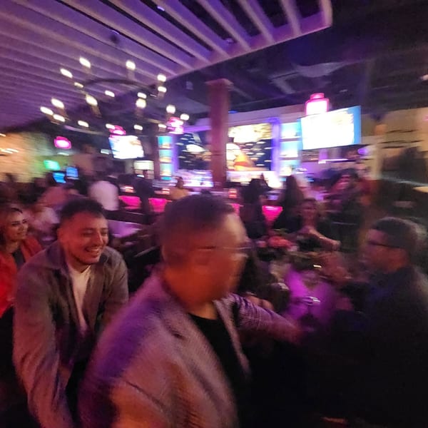 A lively restaurant interior with patrons dining under purple and pink lighting, featuring multiple large screens.