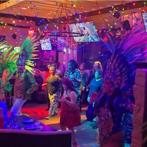 Performers in vibrant feathered costumes entertain guests in a lively, colorful restaurant with slot machines.