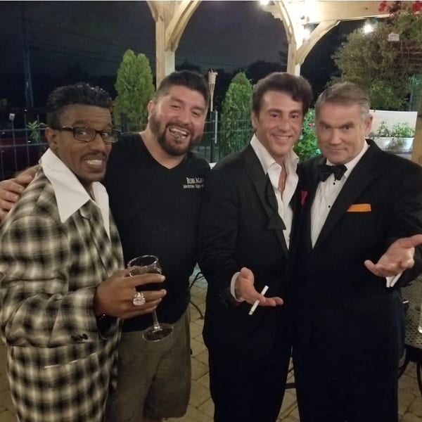 Four men, two in suits, pose together on an outdoor patio at night.
