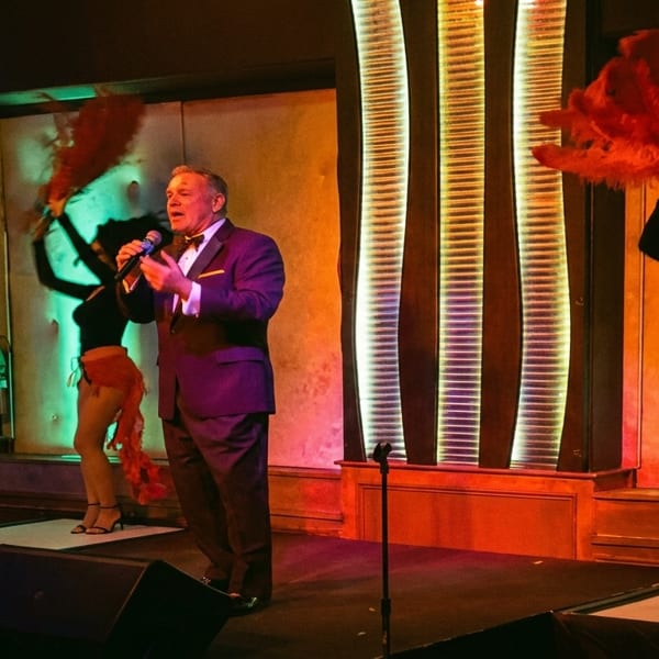 A male singer in a suit performs on stage with two female dancers in burlesque costumes.