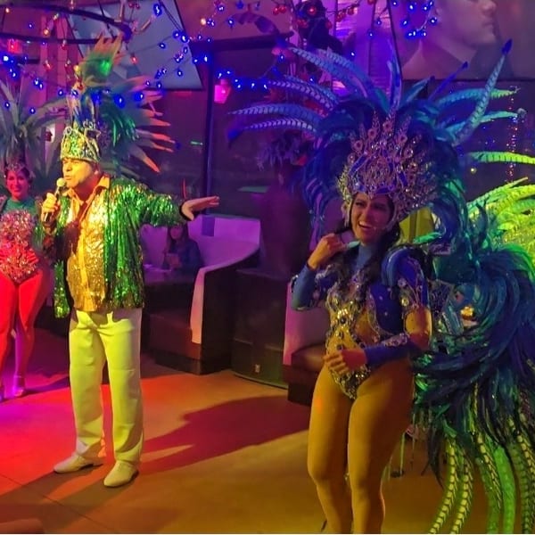 Performers in vibrant feathered costumes and a singer entertain guests in a lively restaurant.