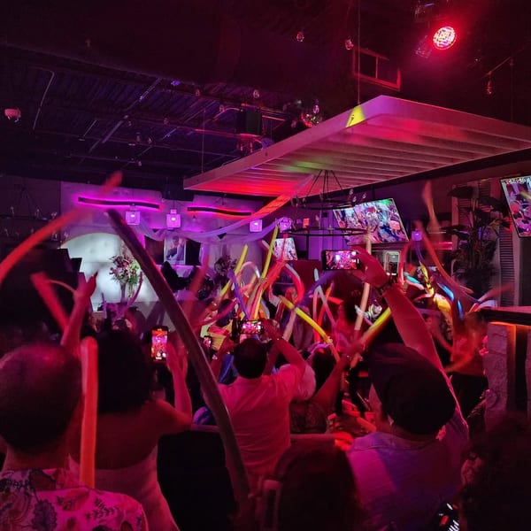 People dancing with colorful balloon animals under vibrant purple and red club lights.