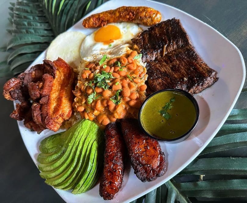 A large platter of Colombian bandeja paisa with steak, beans, rice, avocado, plantains, and fried egg.