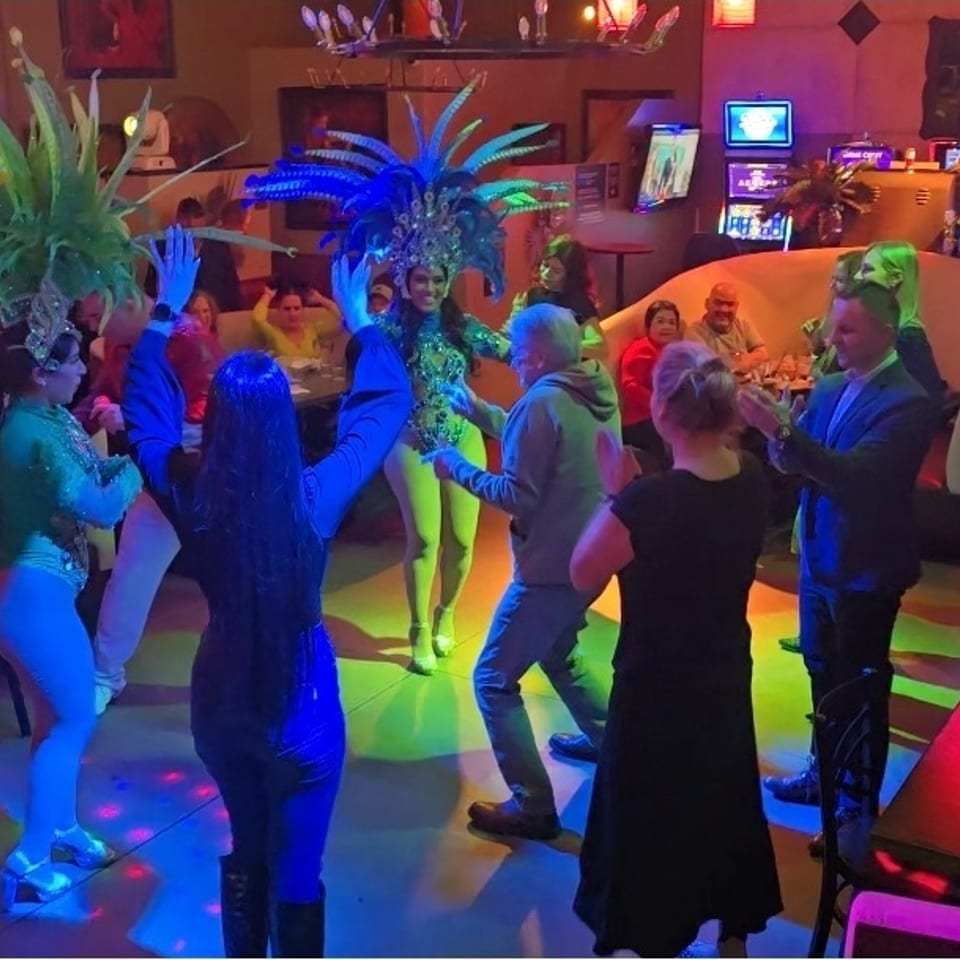 Performers in elaborate costumes dance with guests in a lively, colorful restaurant setting.
